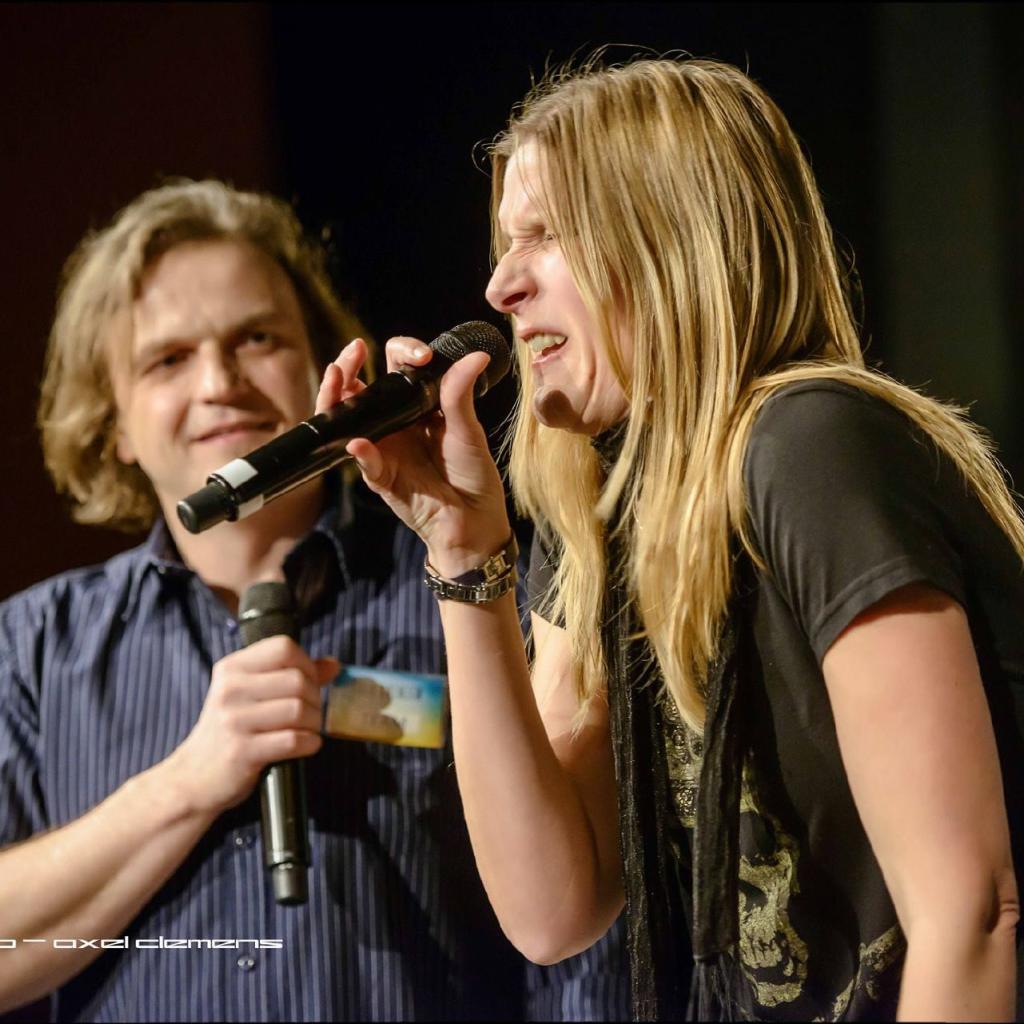 Stephanie und Dirk stehen auf einer Bühne mit Mikrofonen, Stephanie verzieht das Gesicht beim Sprechen