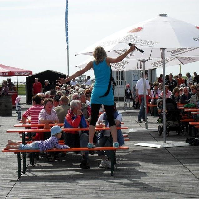 Stephanie steht in St-Peter Ording auf einer Bierbank und animiert die vor ihr sitzenden Gäste