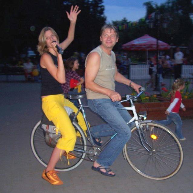 Stephanie singt in ein Mikrofon, während sie von einem Radfahrer auf dem Gepäckträger mitgenommen wird